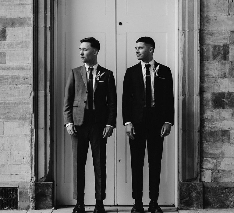 Grooms look to their right together as they stand in front of white doorway in suits