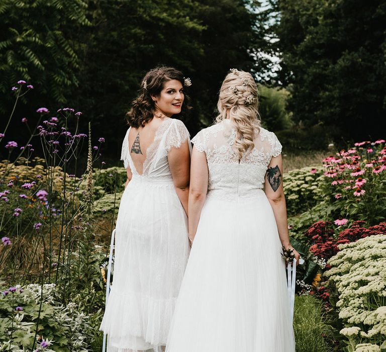 Bride with brunette hair looks back at the camera as she walks with bride with blonde hair through the gardens of Chaucer Barn venue