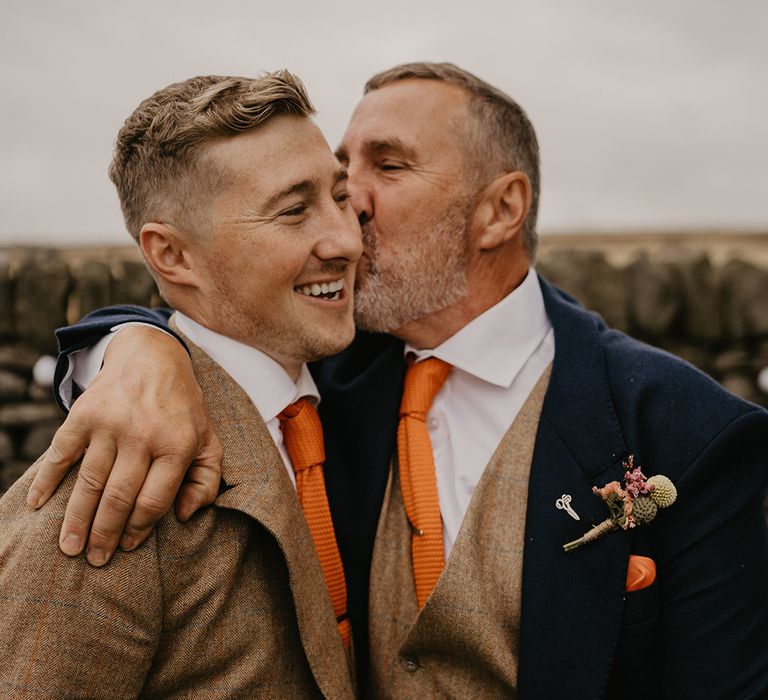 Father in dark suit jacket and brown waistcoat kisses groom on the cheek in matching brown suit and orange tie