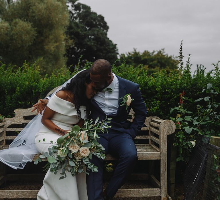 Groom in blue suit and mint green bow tie puts his arm around bride as they sit on bench together 