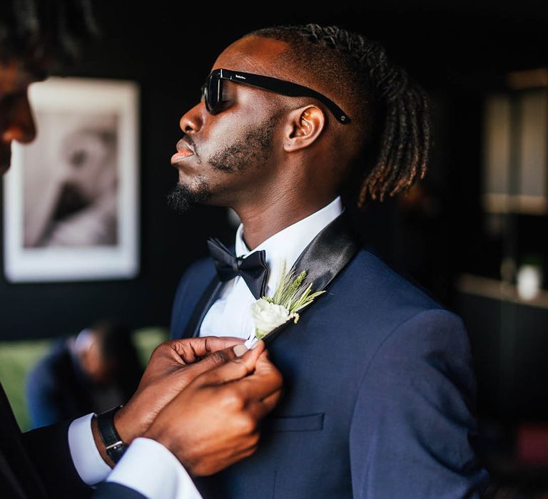 Groomsmen in blue and black tuxedo gets his white flower buttonhole attached 