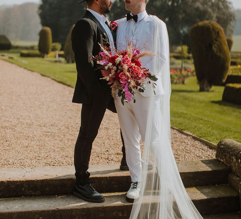 Groom in a black suit and fedora hat standing with his partner at Prestwold Hall Barns in a white groom suit and cape holding a vibrant red bouquet 