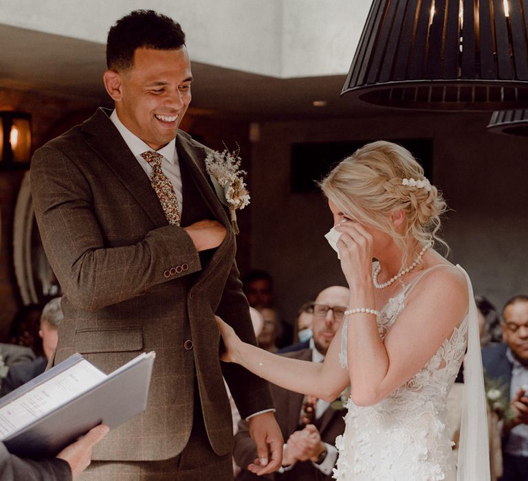 Bride in 3D lace wedding dress tips her happy tears with groom in brown checkered suit and patterned tie