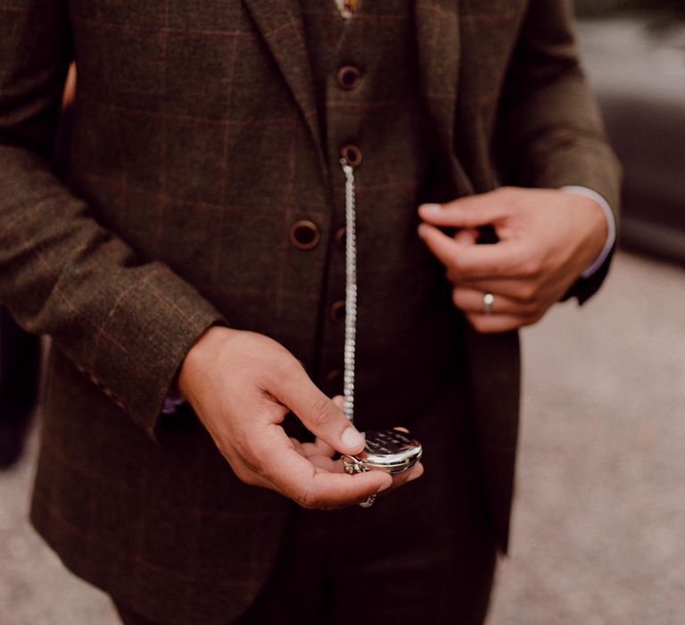 Groom in three piece brown checkered suit with dried buttonhole gets out his silver engraved pocket watch