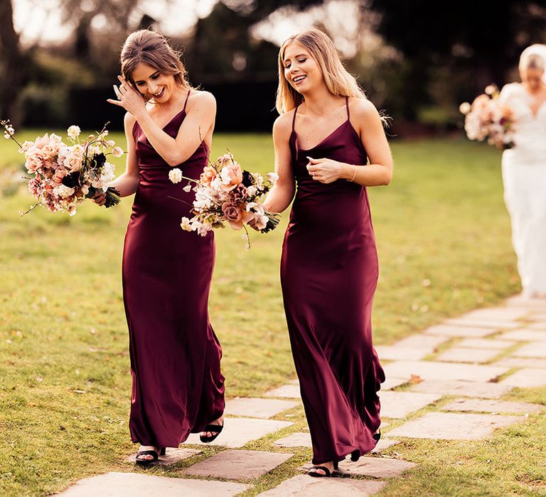 Bridesmaids in burgundy satin cowl neck bridesmaid dresses walk in front of the bride and father of the bride as they walk down the aisle 