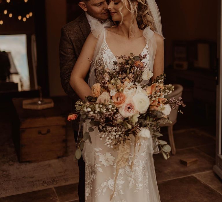 Bride in Made With Love lace wedding dress with bow sleeves holding autumnal wedding bouquet being hugged from behind by the groom in brown suit for rustic wedding 