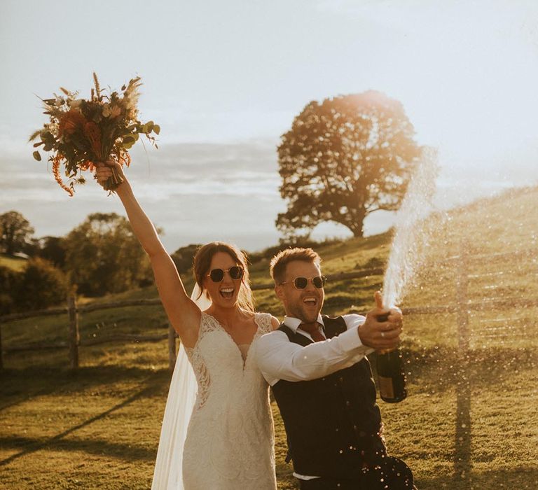 Bride in lace dress with long veil and neutral bouquet with groom doing champagne pop