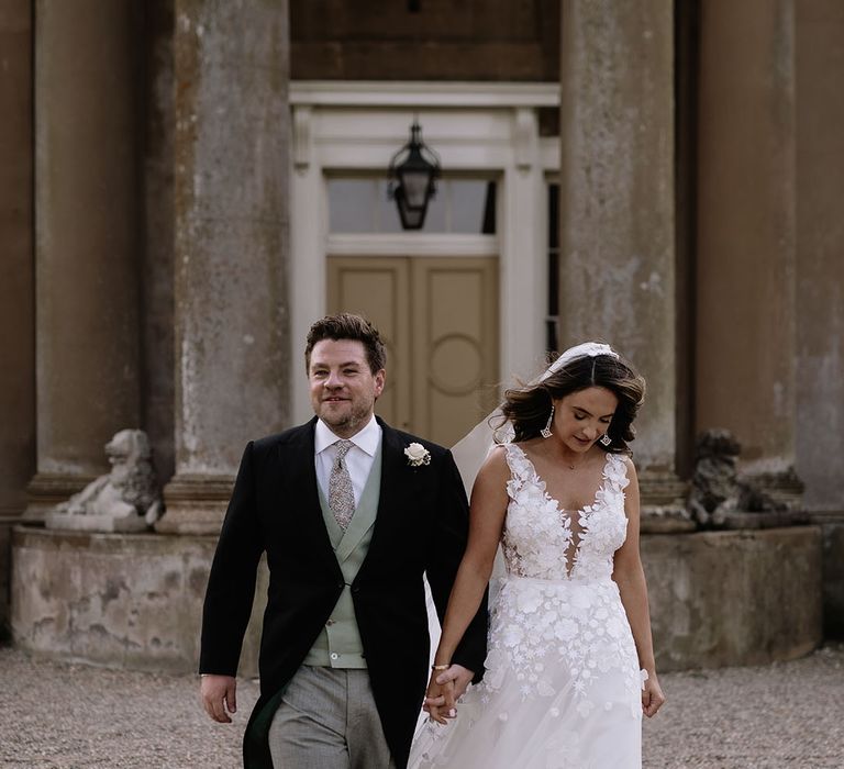 Bride in floral applique wedding dress walks around holding hands with the groom in morning suit with green waistcoat 