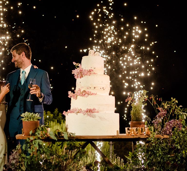 Bride & groom stand in front of rustic four tier wedding cake as sparklers light behind them in the night sky 