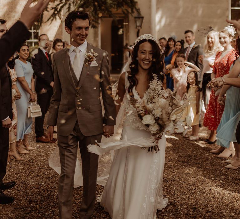 Bride wearing a bridal headband carrying a white flower dried grass bouquet and groom in grey suit have confetti exit from wedding 