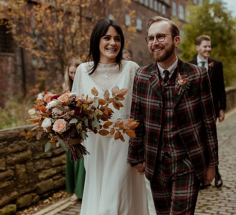 Bride carries Autumnal floral bouquet and wears lace cape as groom wears bespoke tartan suit 