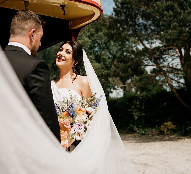 Bride wears a pearl embellished veil holding a bright and colourful wedding bouquet as she gazes at the groom 