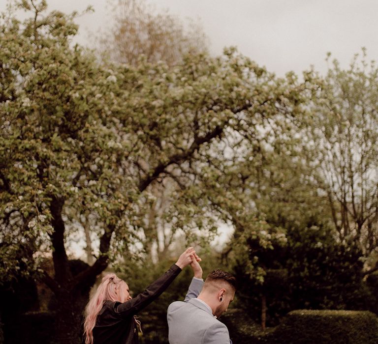 Bride with pink hair wears black denim jacket outdoors as she walks with her groom in pale grey suit 