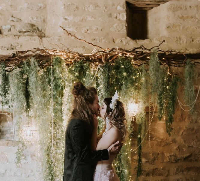 Groom in a tuxedo and bride in a pink strapless wedding dress kissing at the altar with a twig, fairy light and foliage backdrop 