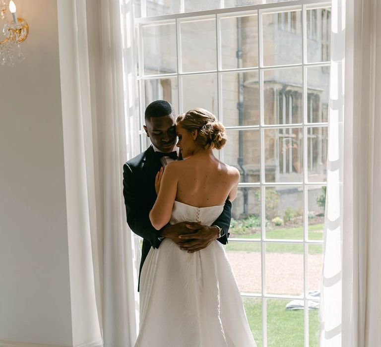 Bride in strapless and pleated brocade Jesus Peiro wedding dress hugging the groom as they stand next to a window 