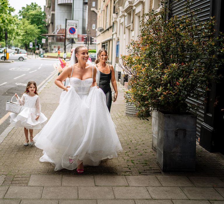 Bride wears sparkly wedding dress and walks alongside her bridesmaid in silk black bridesmaid dress and flower girl carrying white wicker basket