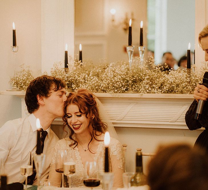 Groom kisses his bride who wears her red hair in curled half-up half-down styling 