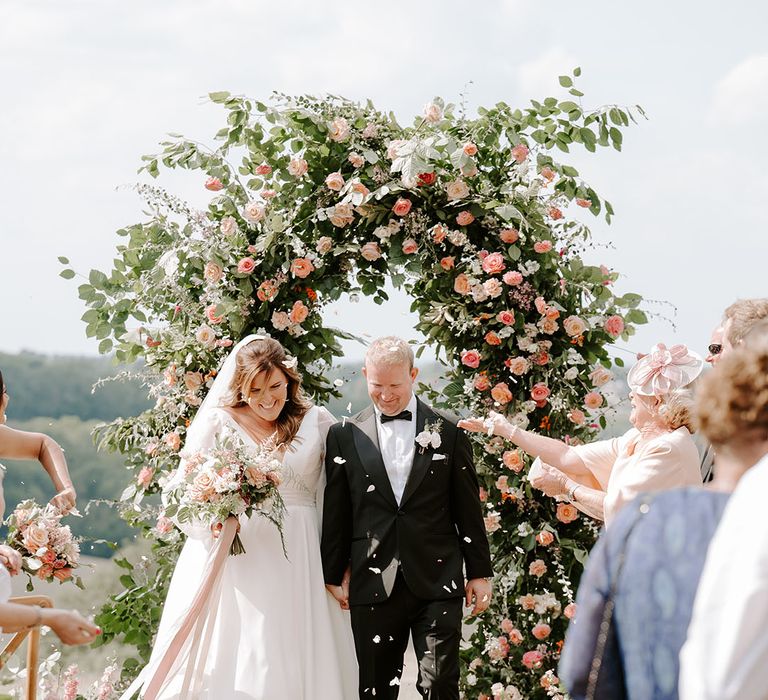 Groom in black-tie walks through white petals with his groom after outdoor wedding ceremony with floral arch