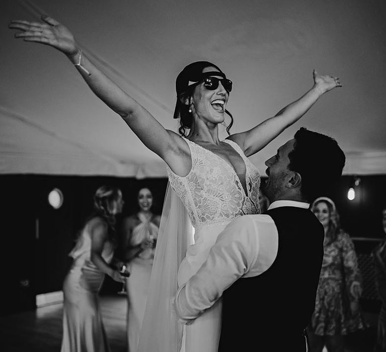 The groom lifts the bride while she wears a cap backwards and some sunglasses