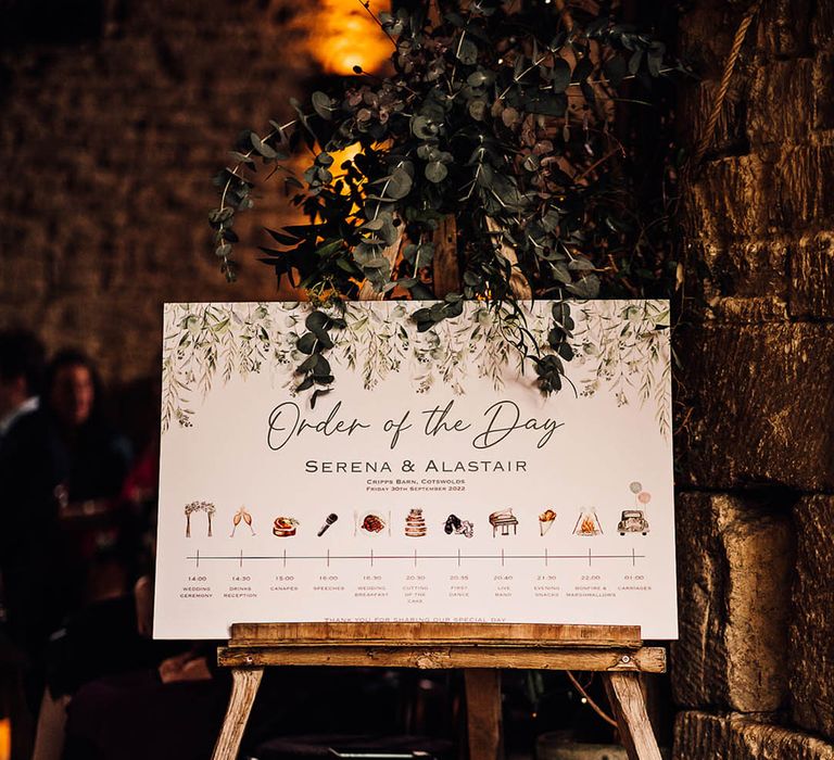 A wedding order of the day sign with images of each part of the day and foliage decor 