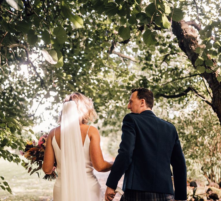 Groom holds bride's wedding dress train as they walk together after wedding ceremony