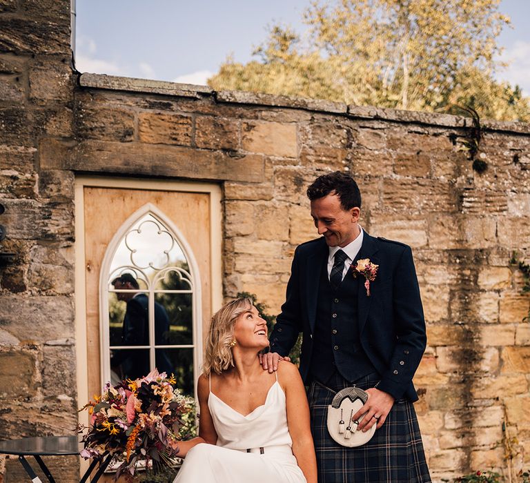 Groom in traditional Scottish kilt looks lovingly toward his bride who wears silk slip wedding dress