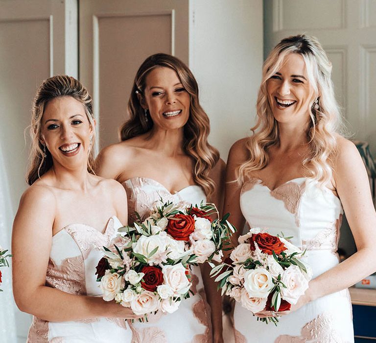 Bridesmaids in white strapless sweetheart dresses with rose gold pattern holding bouquets of roses 