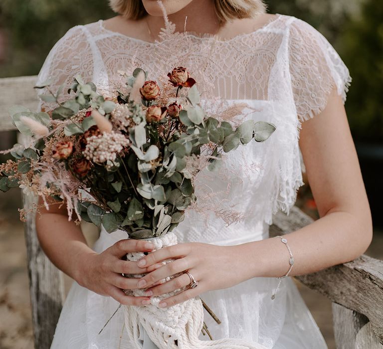 Bride wearing elegant jewellery in a sheer lace boho wedding dress holding a matching bouquet and dried flower crown 