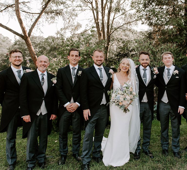 Bride in Made With Love wedding dress and flower applique veil with the groom, groomsmen and family members in three piece suits with grey waistcoats and blue ties with clips