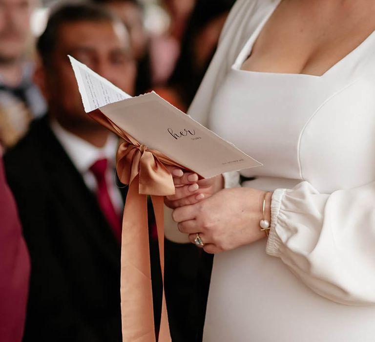 Bride wearing square neck wedding dress with gold and pearl jewellery and pink nails hold her wedding vows tied with orange ribbon 