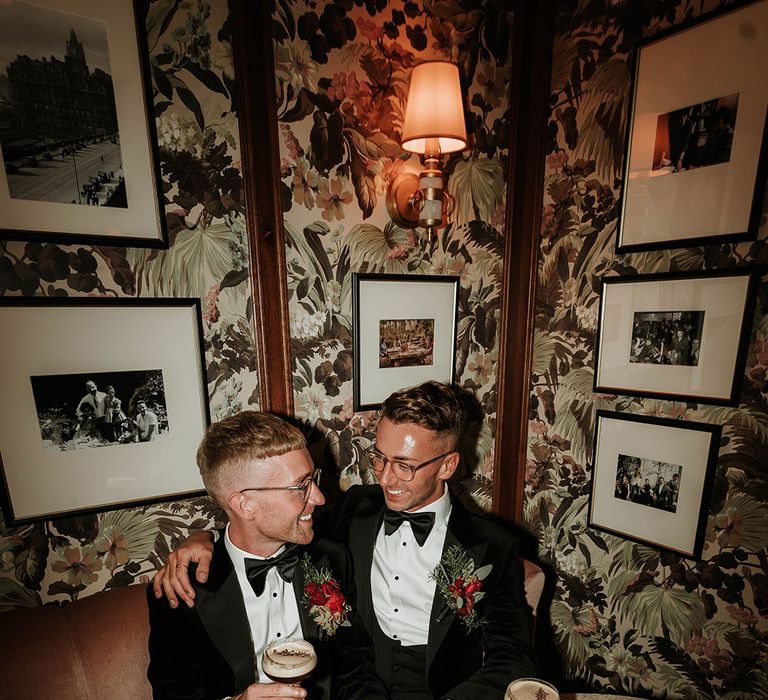 Grooms in matching black velvet suits with bowties and red rose boutonnieres sharing espresso martinis at hotel wedding venue after stylish, contemporary city wedding