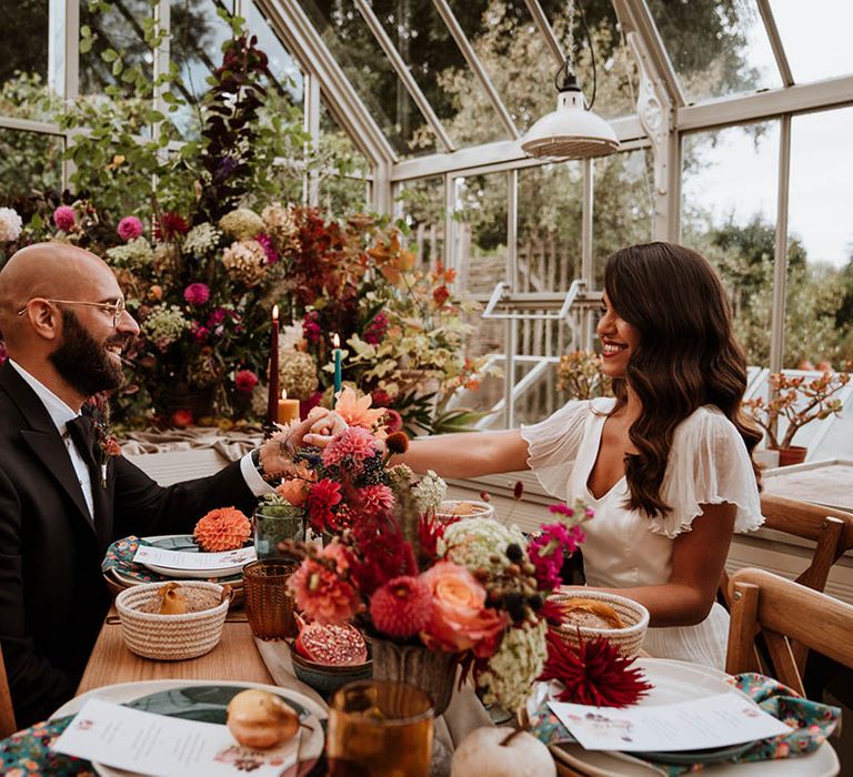 Bride and groom holding hands over neutral, botanical and autumnal wedding tablescape with neutral-toned tablerunner on rustic wooden table, orange, green and red coloured glasses, earthenware, seasonal fruit and vegetables, red, brown and green tapered candles in gold candlesticks and clear vases with autumnal coloured flowers