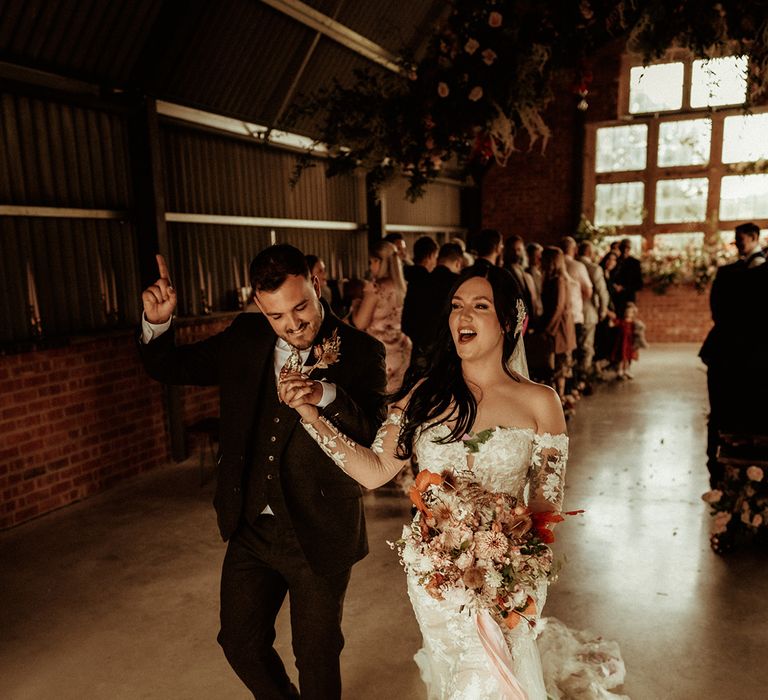 Newlyweds dancing; groom in a dark suit with flower patterned tie and pocket square and dried flower boutonniere and bride in off the shoulder bodycon wedding dress with long, sheer sleeves and lace detailing holding large dried flower bouquet with baby pink ribbon