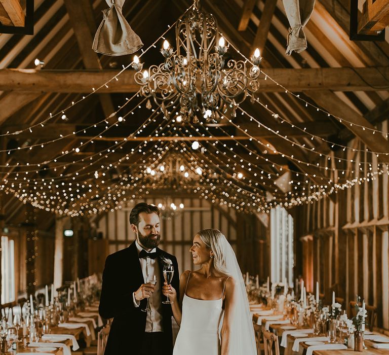 The bride in a fitted wedding gown with thin straps, gold hoops and a veil smiles at the groom as they share champagne in their fiery lit wedding venue 