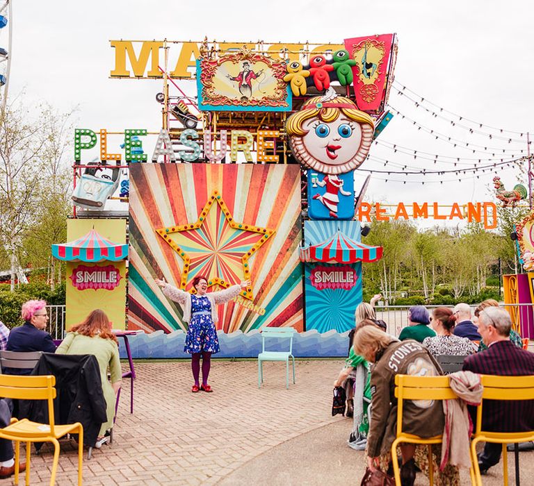 Wedding celebrant wearing blue patterned dress and cover up giving speech at theme park wedding with rides, rollercoasters and Ferris wheels in the back 