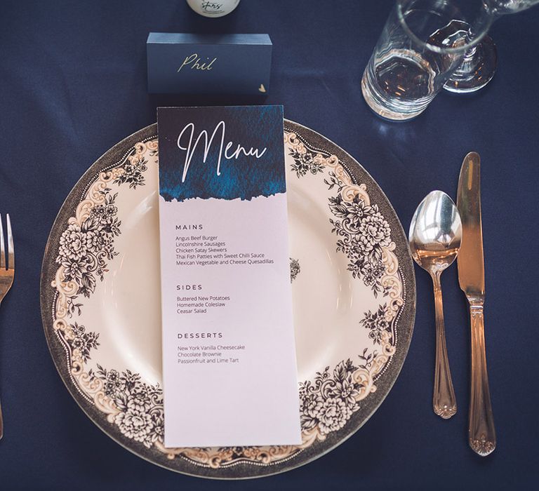 Black and white wedding plate with floral pattern with blue and white wedding menu with silver cutlery and navy blue place name 