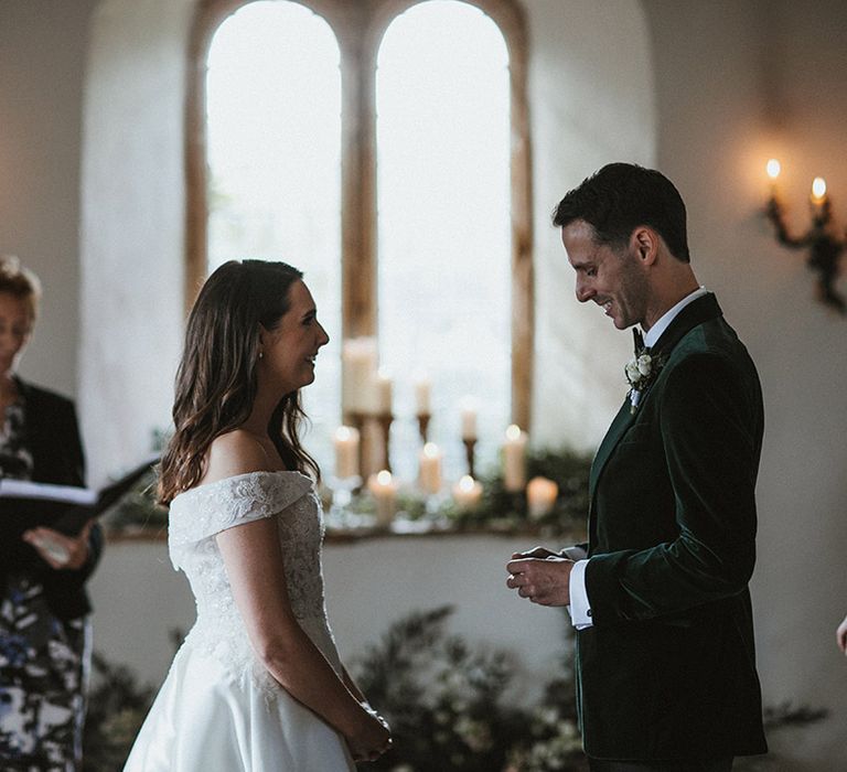 Groom in a dark green velvet tuxedo smiles as he reads out his wedding vows to the bride standing opposite him in an off the shoulder lace wedding dress