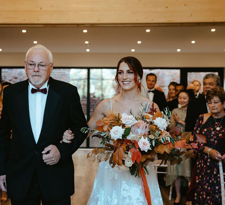 Bride holding burnt orange bridal bouquet walks down the aisle with her father in black tie 