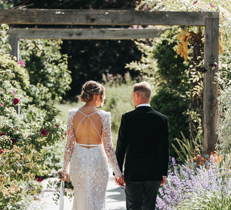 Bride with her hair in a chic updo with pearl hair pins in an open back wedding dress with lace detail walking with the groom in a black tux for their traditional wedding 