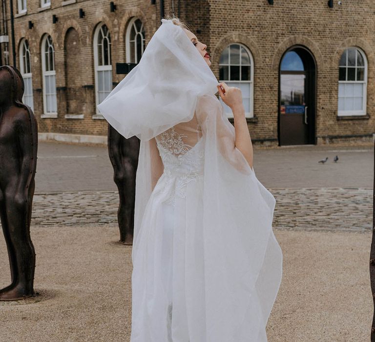 Bride in sculpted bandeau maxi wedding dress and white sheer wedding cape with hood and dramatic sleeves standing outside London wedding venue 