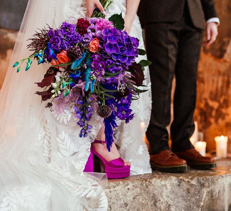 Bride in applique sheer wedding dress holding colourful purple bridal bouquet and wearing open-toe purple platform wedding shoes standing with groom in tan Dr Martens shoes 