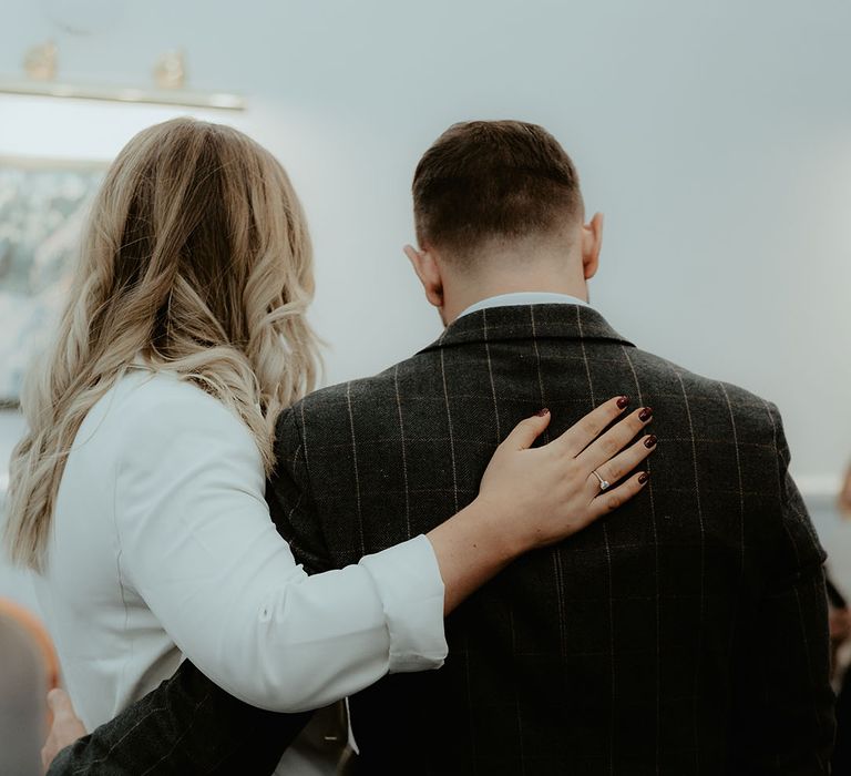 The bride in a white blazer with dark red wading nails wearing her engagement ring stands with her arm around the groom in dark grey suit 