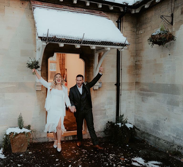 The bride in a split leg wedding dress with ruffle detail holding white flower bouquet in the air with the groom as they exit their wedding ceremony