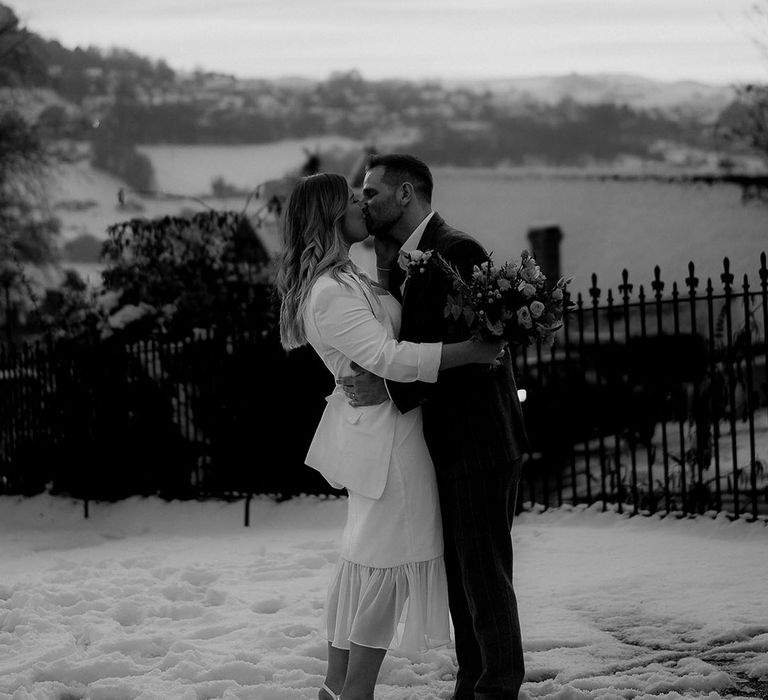 Bride in white dress with ruffle bottom detail holding white blazer and white shoes kissing the groom for winter registry office wedding 