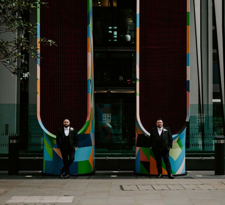 Couples wedding photoshoot at LGBTQ+ city wedding wearing complimenting three piece black tuxedos with off white bow ties, white garden rose, lavender twig and dried foliage boutonniere and grooms Louboutins shoes 