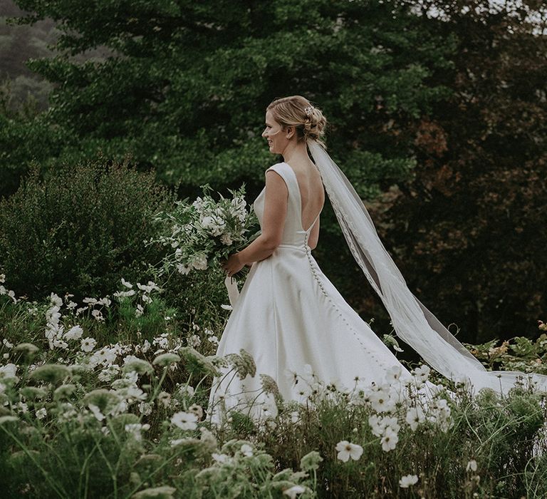 The bride walks through the garden full of white flowers for the wedding in a classic wedding dress with button back detail and embroidered veil 