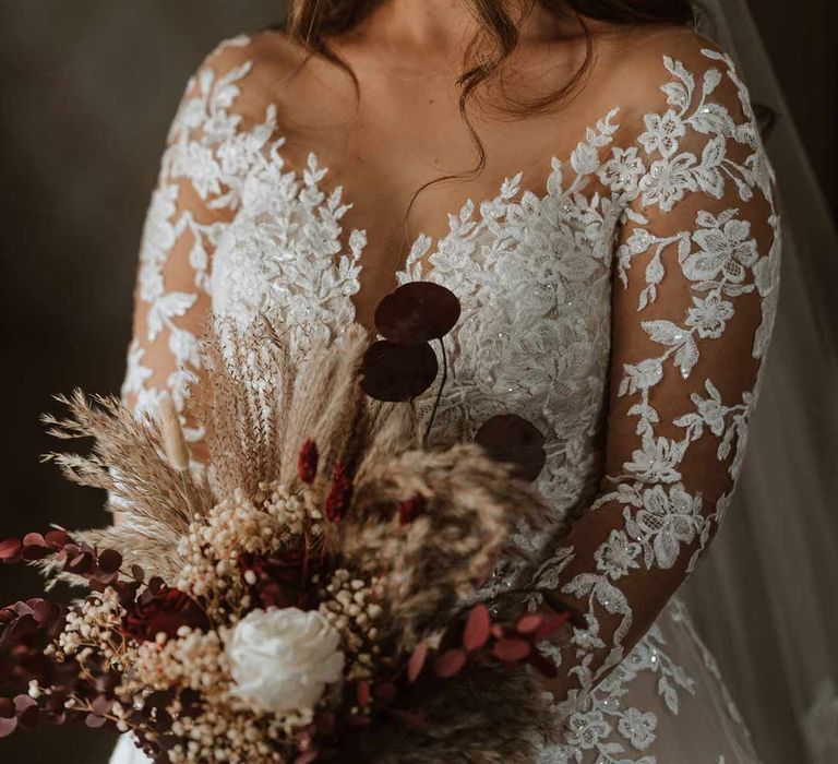 Bride wearing long sleeve lace illusion sleeve wedding dress with overlay holding large autumnal boho dried flower bridal bouquet 