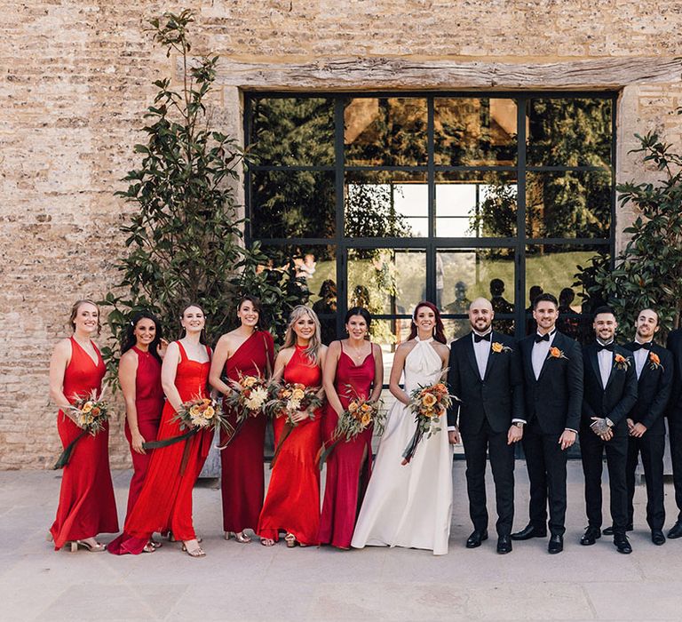 Bridesmaids in satin red bridesmaid dresses with the groom and groomsmen in black tie with the bride in a Halfpenny London wedding dress