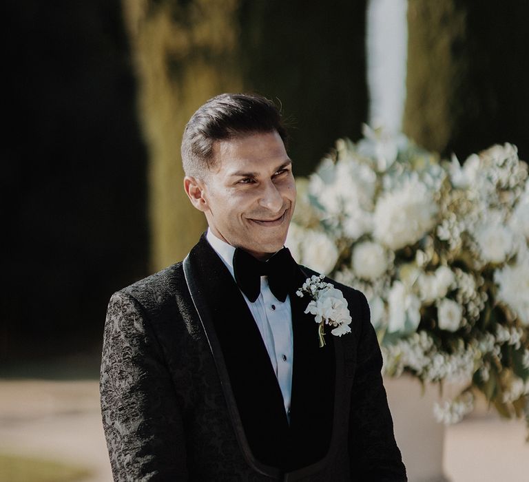 Groom in black tuxedo with black bow tie and white carnation and dried flower boutonniere smiling at bride 