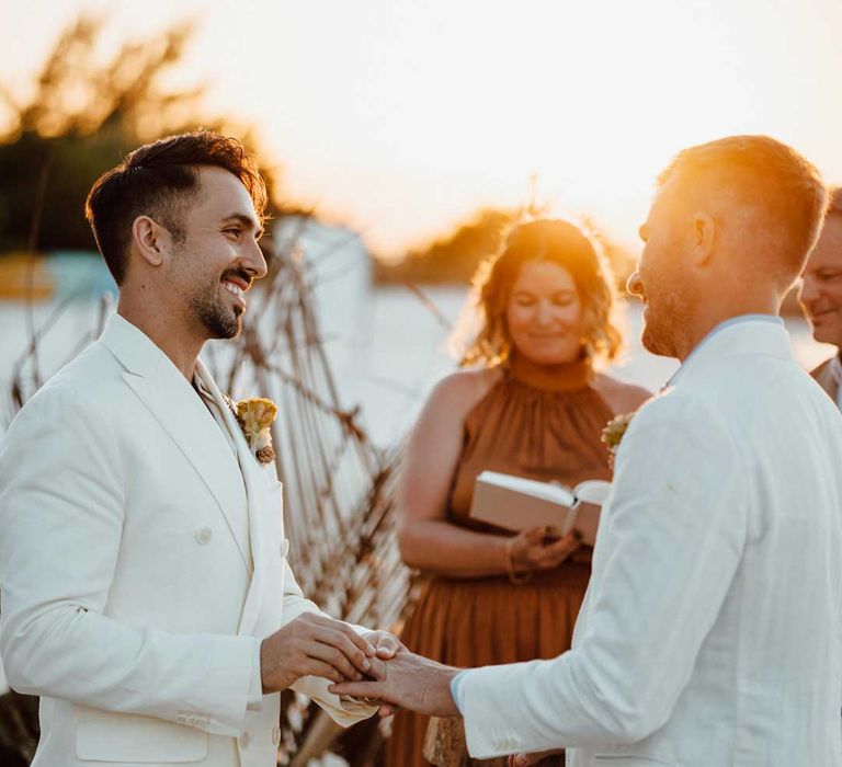 Grooms in complimenting white wedding suits exchanging rings at the aisle on the beach at Mexico destination wedding 